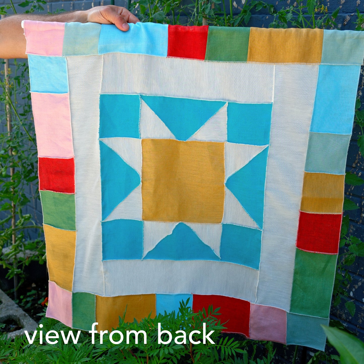 Colorful quilt with star pattern held by a person, view from the back, with greenery in the background.
